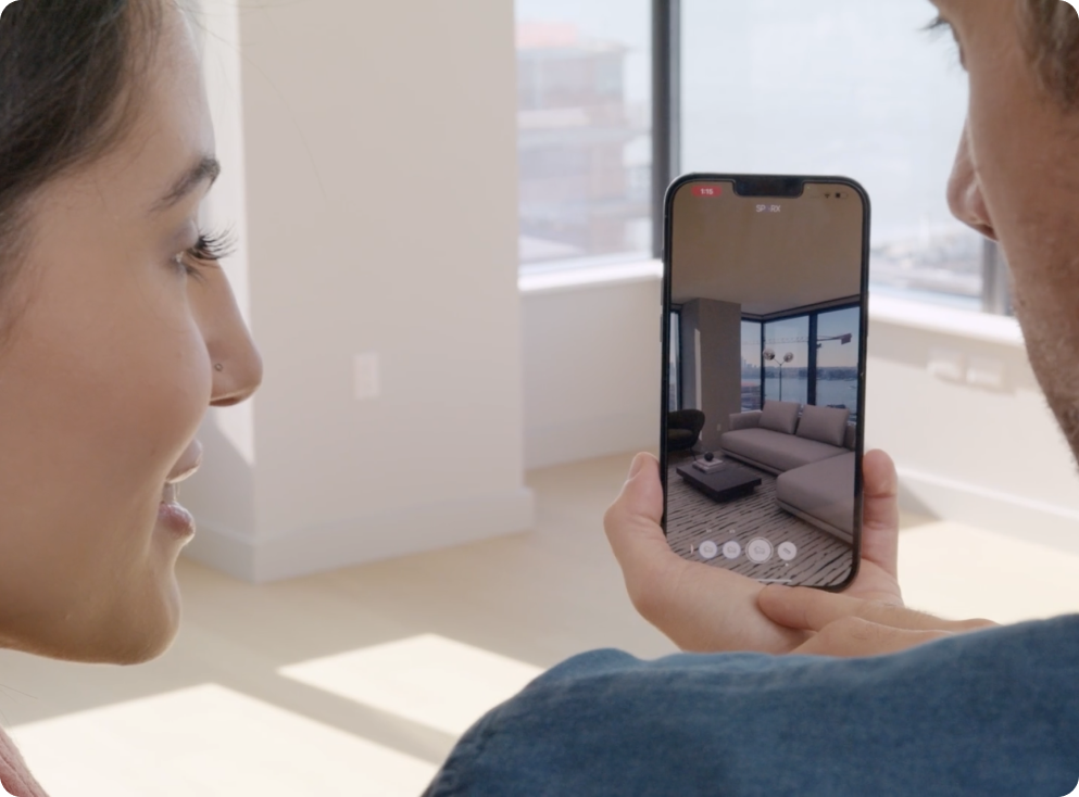 This image shows an empty apartment and the face of a man and woman using augmented reality on an iPhone. On the iPhone's screen is a couch, rug, and table illustrating what the empty apartment could look like.